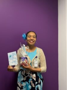 Smiling woman with a blue flower in her hair, turquoise top and cardigan, holding a wrapped gift and a card against a purple wall.